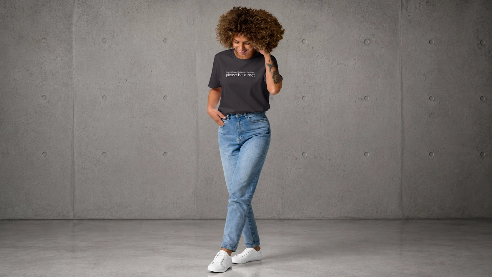 Woman wearing a please be direct t-shirt standing against a gray concrete wall.